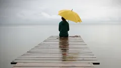 Osamělá osoba se žlutým deštníkem sedí na molu u jezera za deštivého počasí.