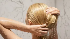 Žena si myje vlasy ve sprše, kapky vody na blond vlasech, koupelna s bílými kachličkami.