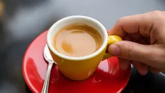 Ruka držící šálek espressa na červeném talířku se lžičkou, káva zblízka.
