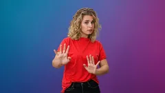 Žena v červeném tričku gestikuluje stop rukama na barevném pozadí