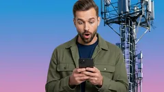 Muž překvapeně sleduje mobilní telefon před mobilní vysílací věží na modrém pozadí.