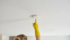 Žena v gumové rukavici ukazuje na mokrý flek a poškození stropu v bytě.