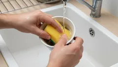 Mytí hrnku žlutou houbičkou pod tekoucí vodou v kuchyňském dřezu.