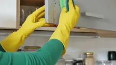 Úklid kuchyně, ruce v rukavicích myjí kuchyňskou skříňku houbičkou.