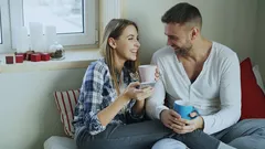 Šťastný pár sedící na posteli, pijící kávu a smějící se ráno doma