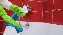 Čištění vany v koupelně, zelené rukavice, sprej a hadřík, červené obklady na pozadí