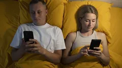 Mladý pár leží v posteli a používá mobilní telefony, žluté povlečení, večer