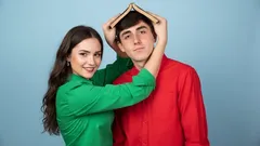 Dívka drží knihu nad hlavou muže, oba se smějí na modrém pozadí.